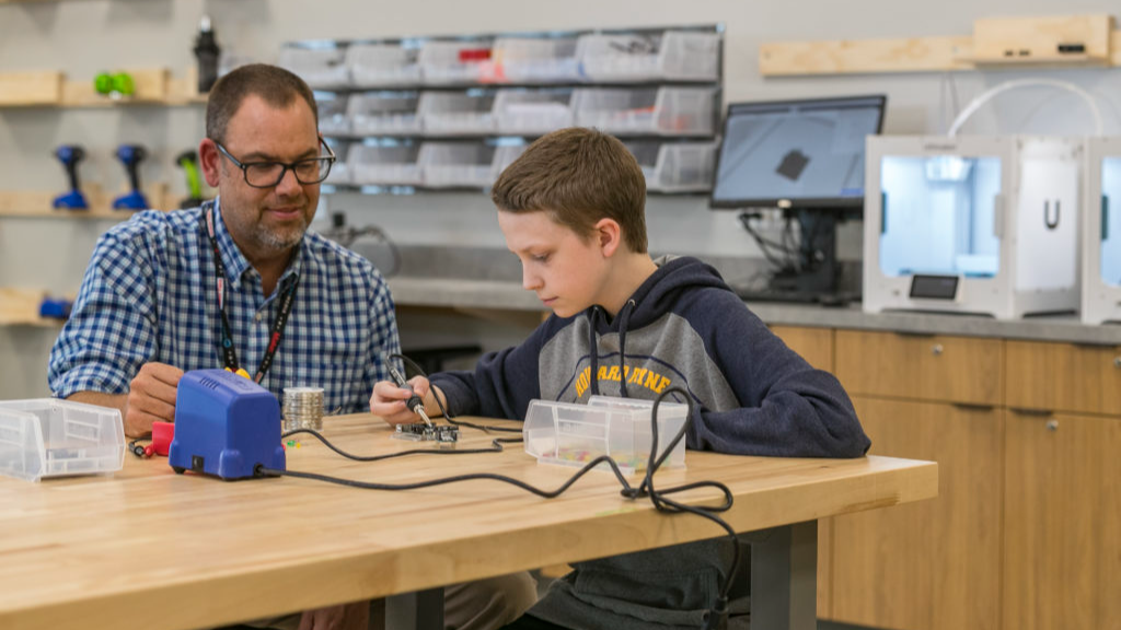 a teacher assisting a student who is welding a piece of circuitry