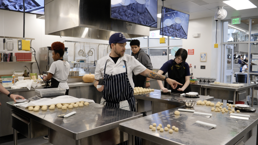 a chef directing students as they work at a metal counter