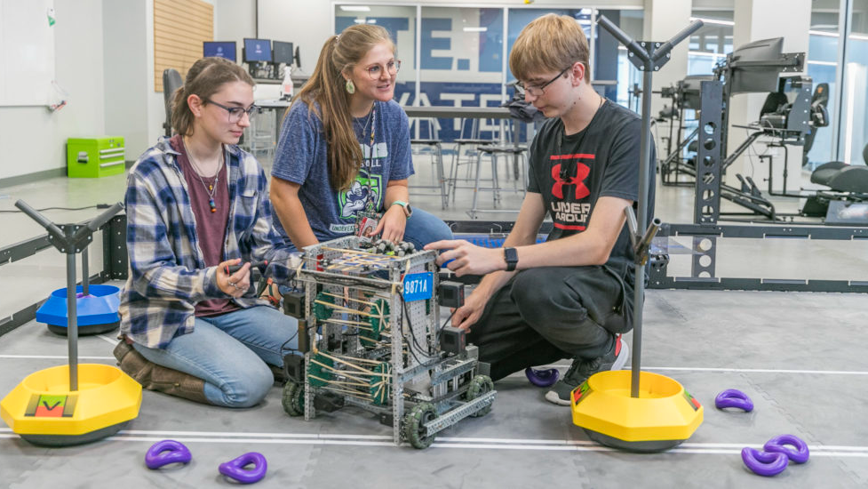 an instructor standing up and speaking to two students who are building a robotic structure