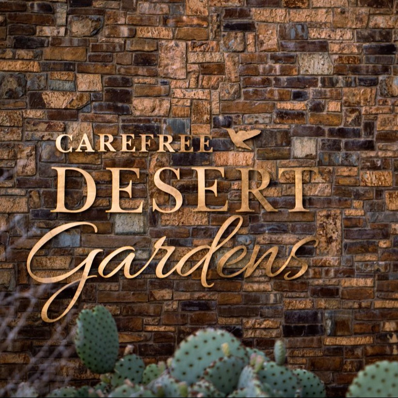 A sign for "Carefree Desert Gardens" on a brick wall with a cactus in front of it.