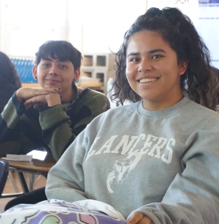 2 students sitting on their desks smiling at the camera