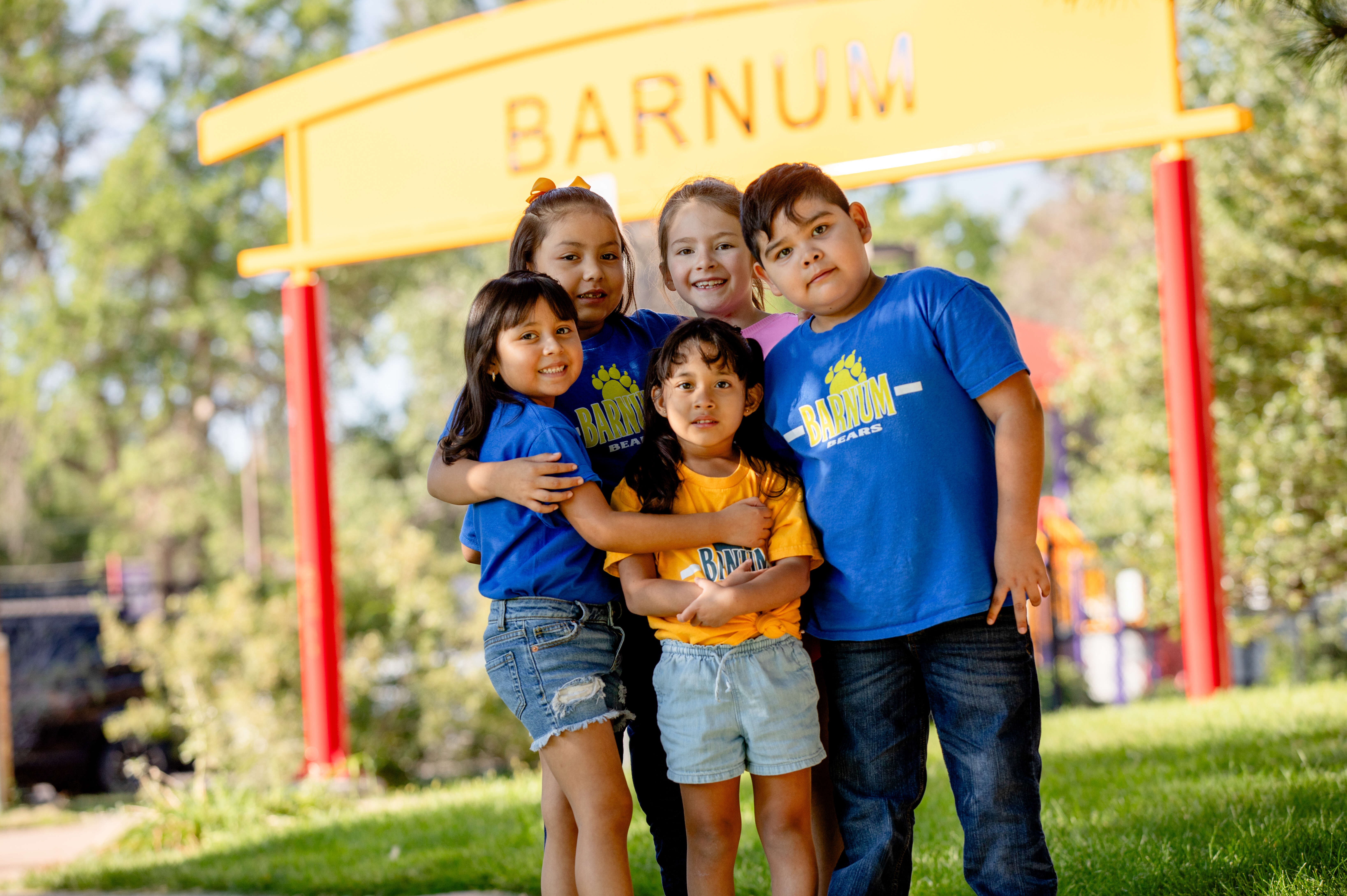 Five children pose together in front of a yellow sign reading "BARNUM". They wear casual summer clothing and are standing in a park.