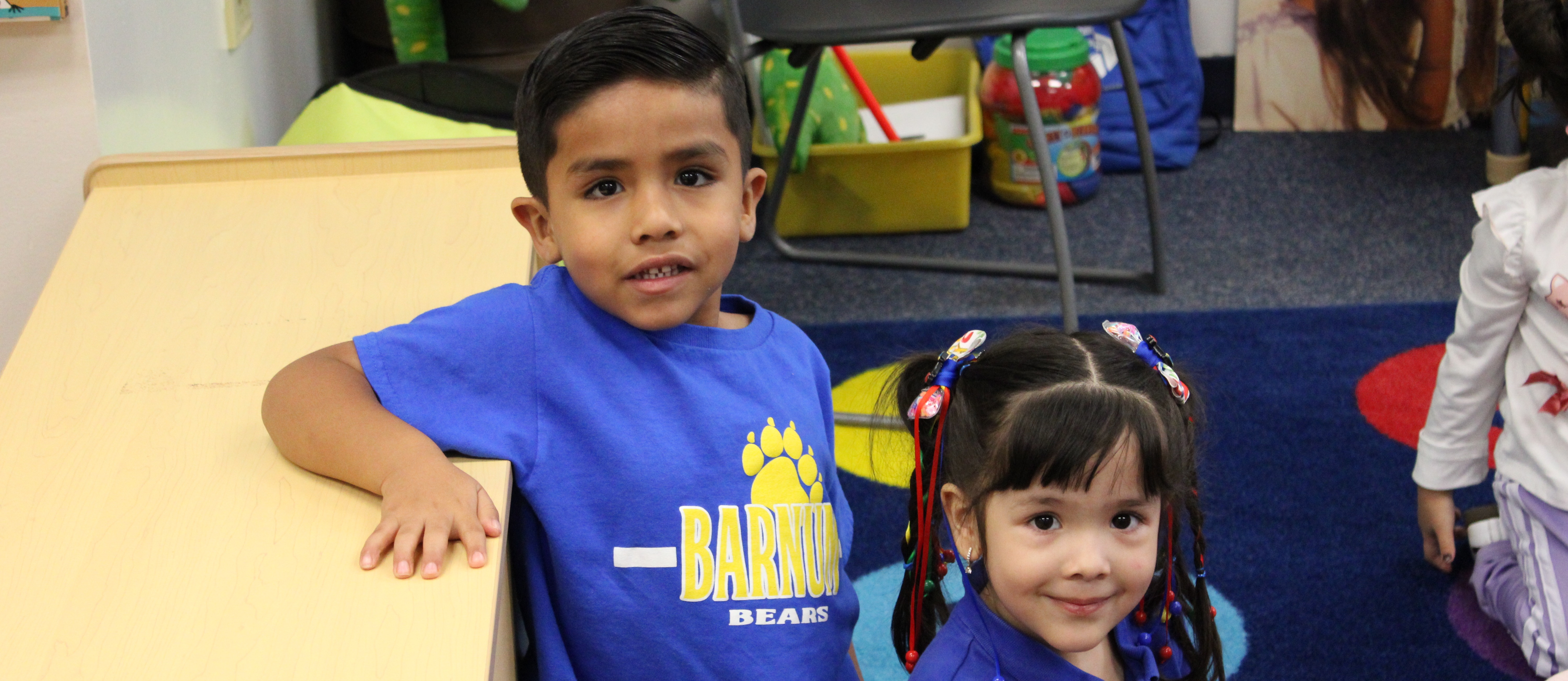 A boy and girl in a classroom. The boy wears a blue shirt and the girl a pink shirt.