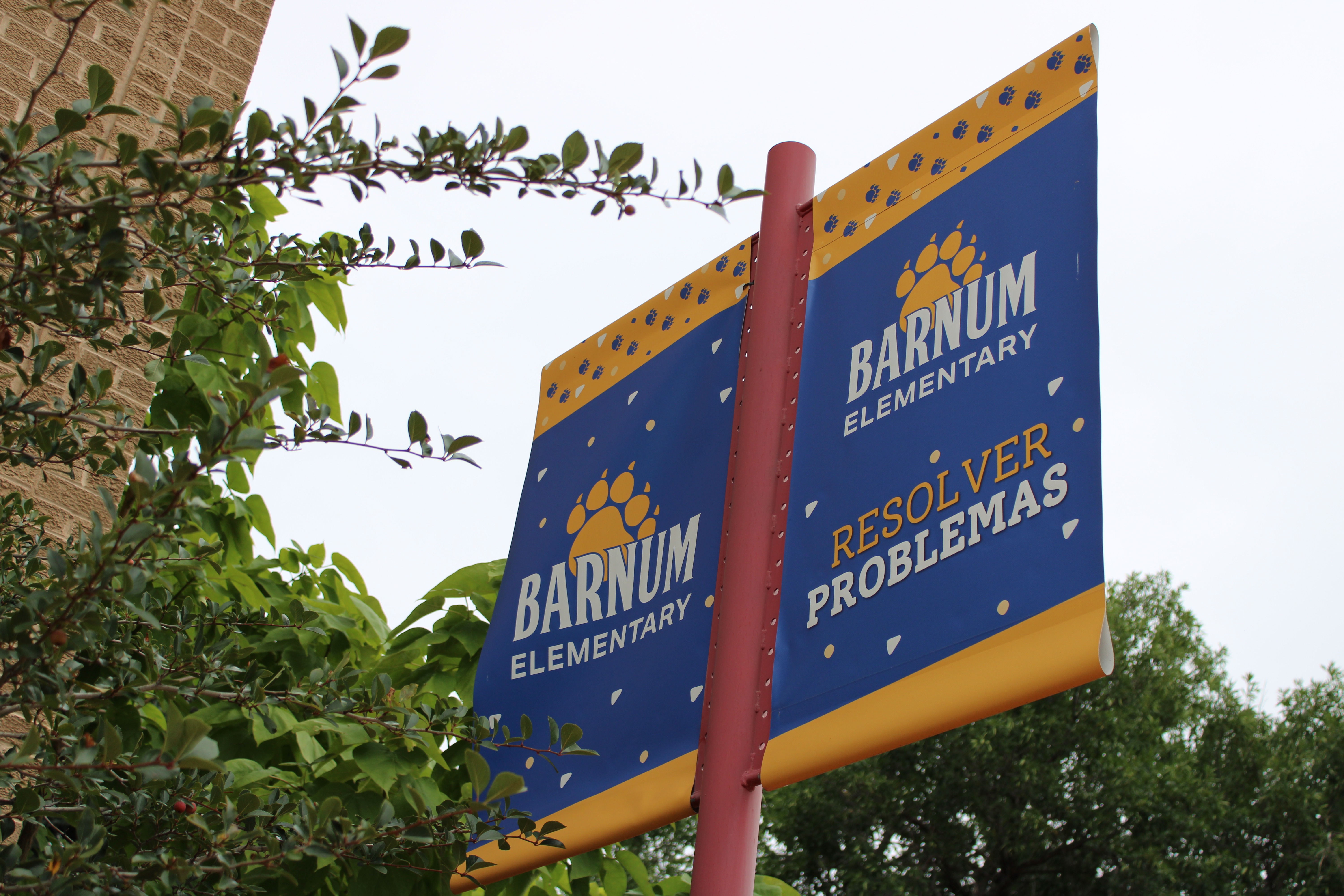 Blue and yellow sign with "Barnum Elementary" and "Resolve Problems" on a pole near a tree.