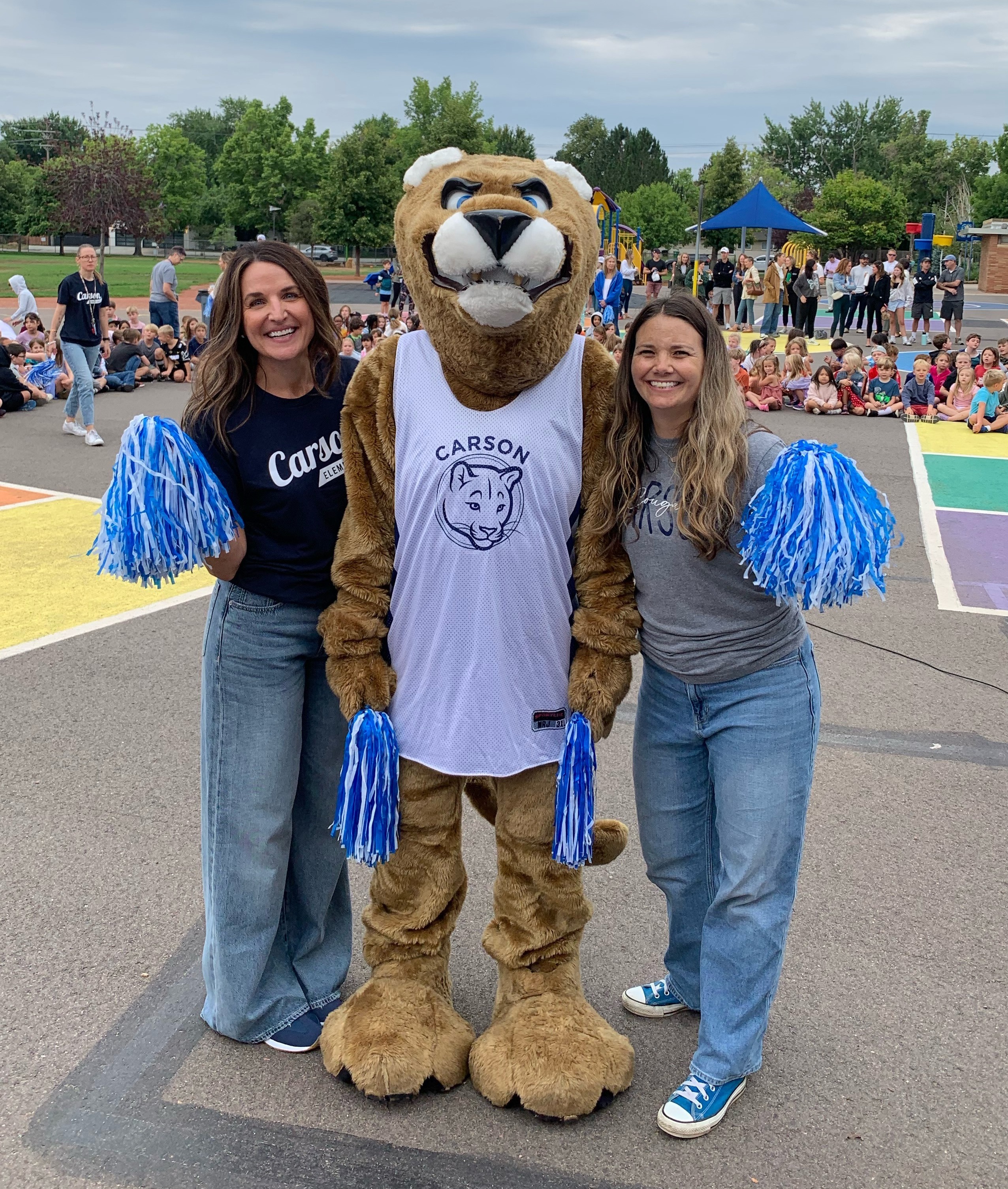 Two women and a mascot stand in an outdoor area with a crowd. The mascot wears a white shirt.