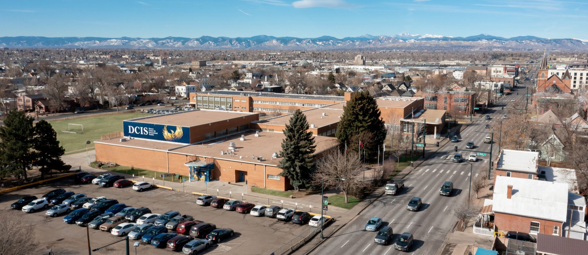 A city street with parked cars. A building with blue signage. Mountains in the distance.