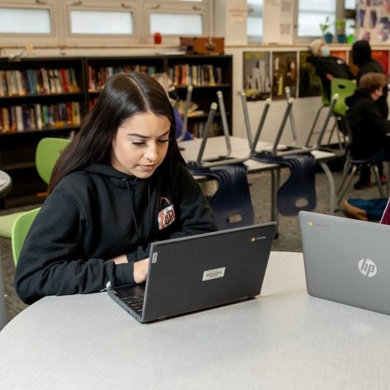 Two children seated in a classroom, using laptops on a table. Bookshelves and other students are in the background.