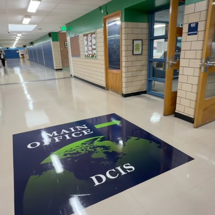 An empty hallway with blue lockers on the left, a floor sign reading "MAIN OFFICE" and "DCIS" on white tiles.