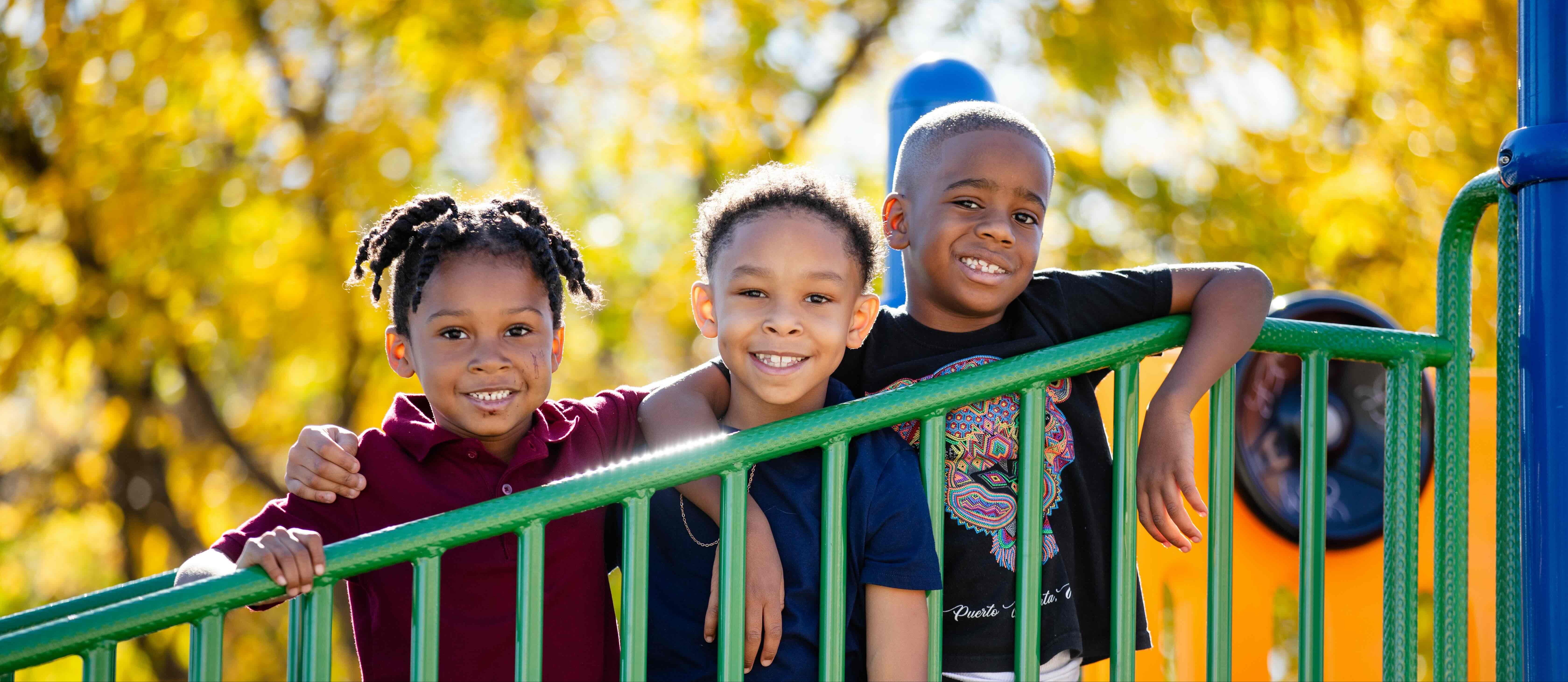 Three children standing in a playground with one holding onto a metal railing.
