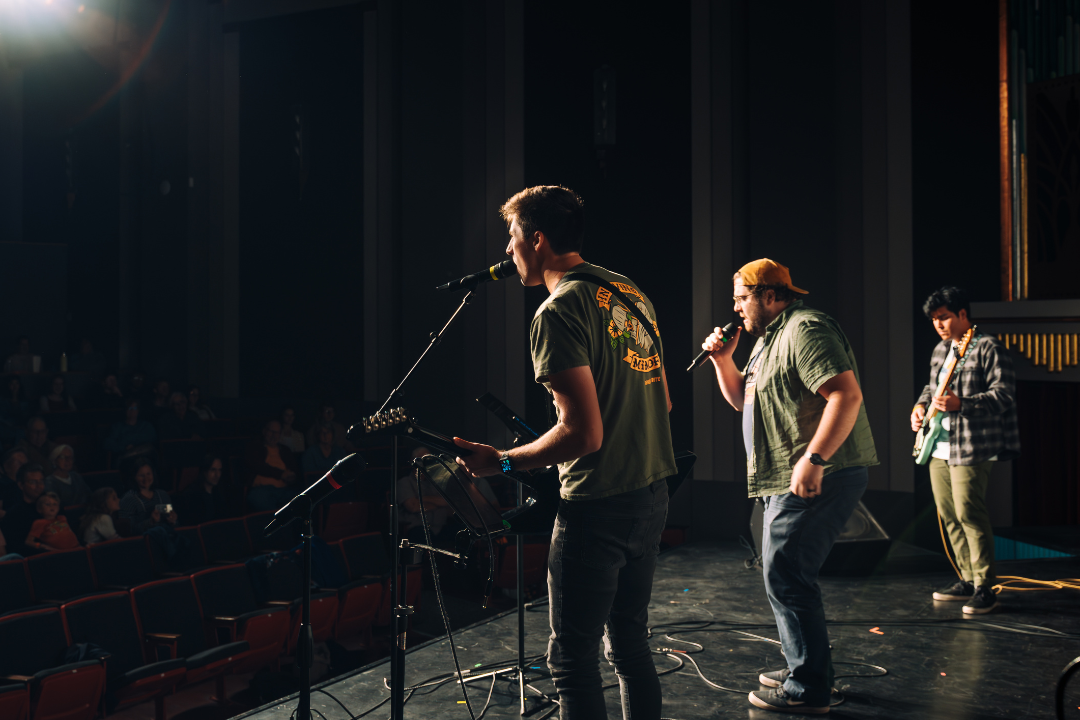 3 men performing on stage, shot from behind, you can see a few rows of the audience in front of the stage enjoying the show at the Romance. Two of the men have guitars, two have microphones. 