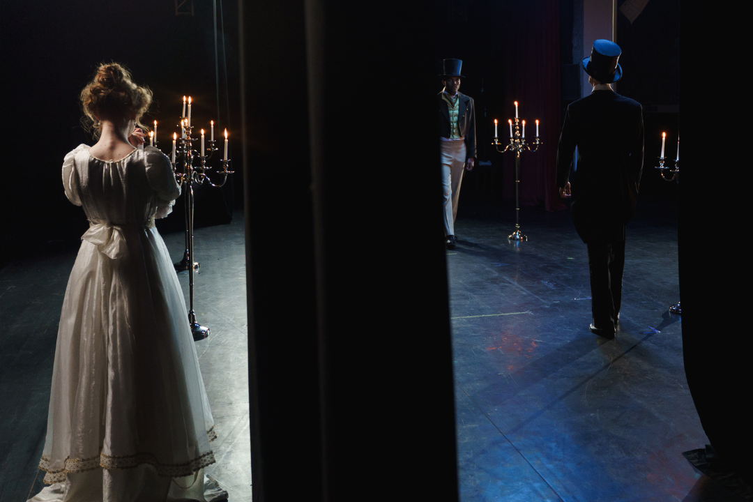 image shot from backstage of 3 actors on stage;  two men in top hats and period-wear facing each other up front and a woman in white period dress with a candleabra a ways behind them watching. 