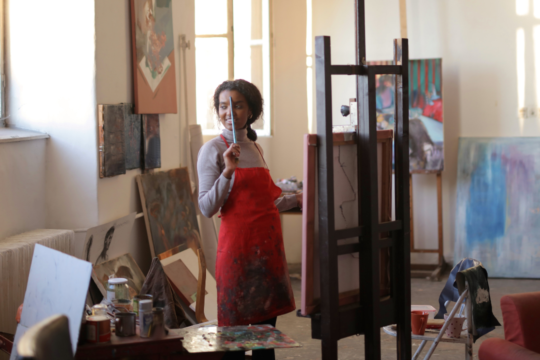 woman with a paintbrush in front of an easel in a studio space with paintings surrounding her