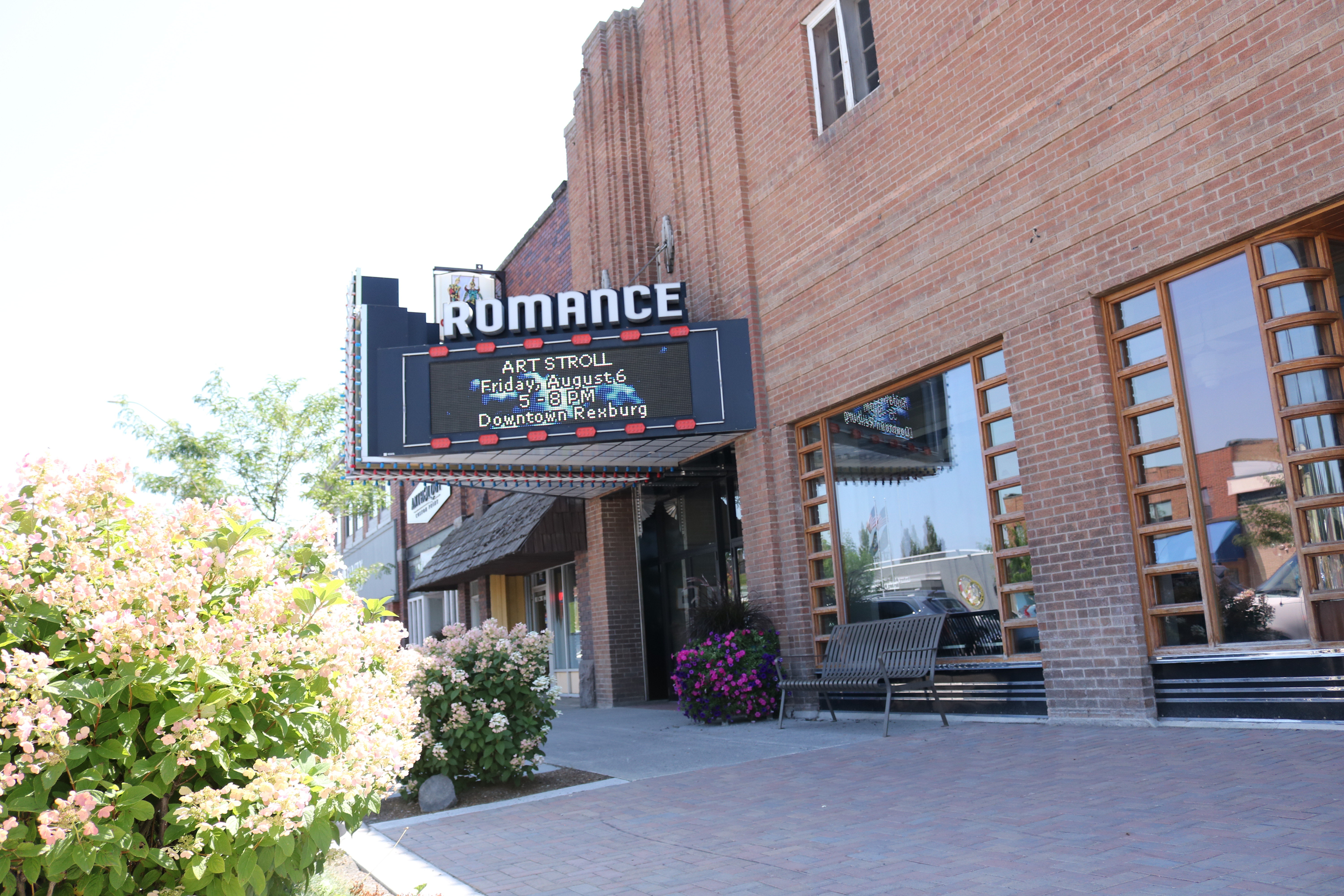 image of the front of the Romance Theater in Rexburg in the summer with bright sunshine and flowers out front. "Art Stroll" advertisement is on the digital marquee screen.