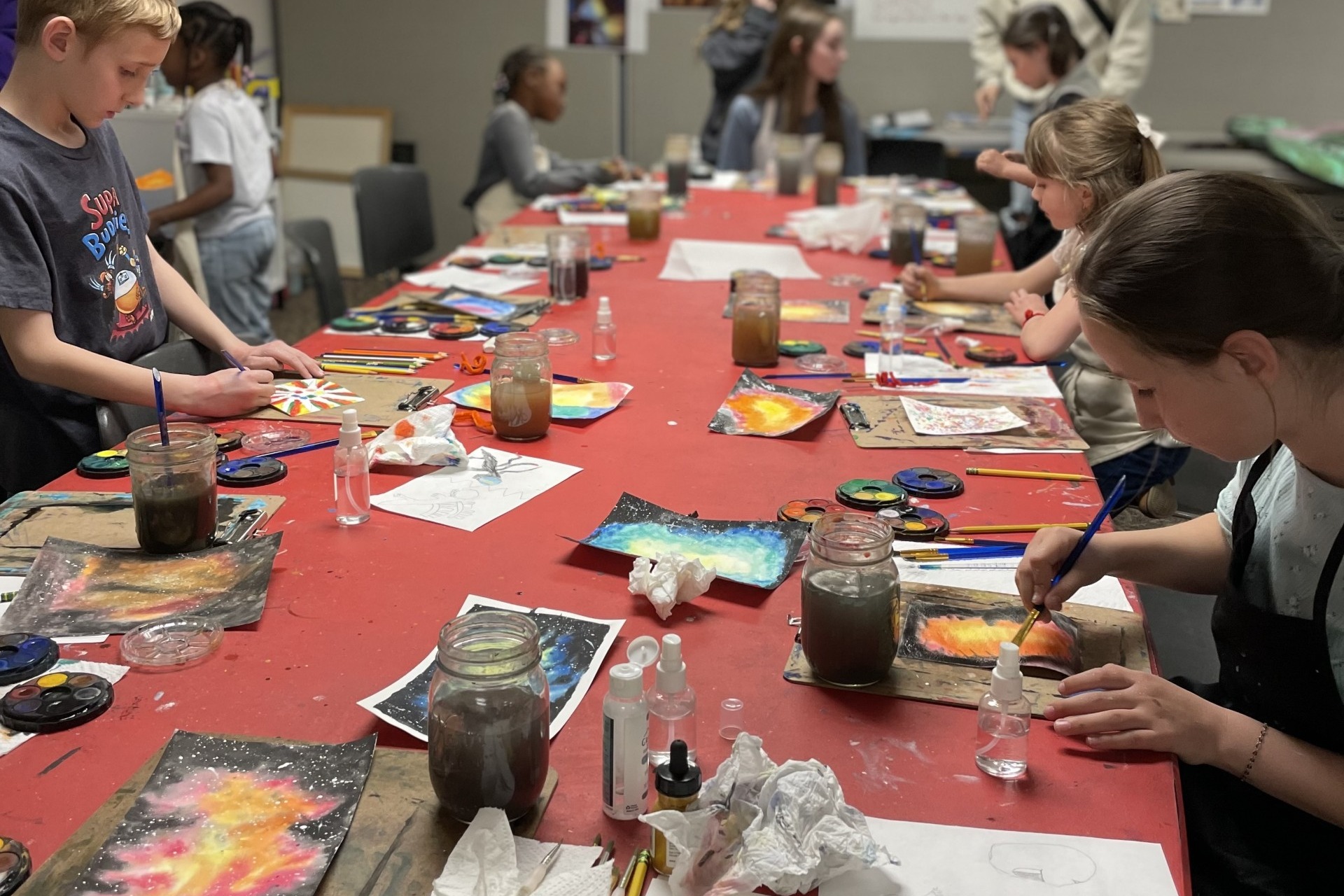 kids painting on a table coverd in red with painting supplies strewn about. 