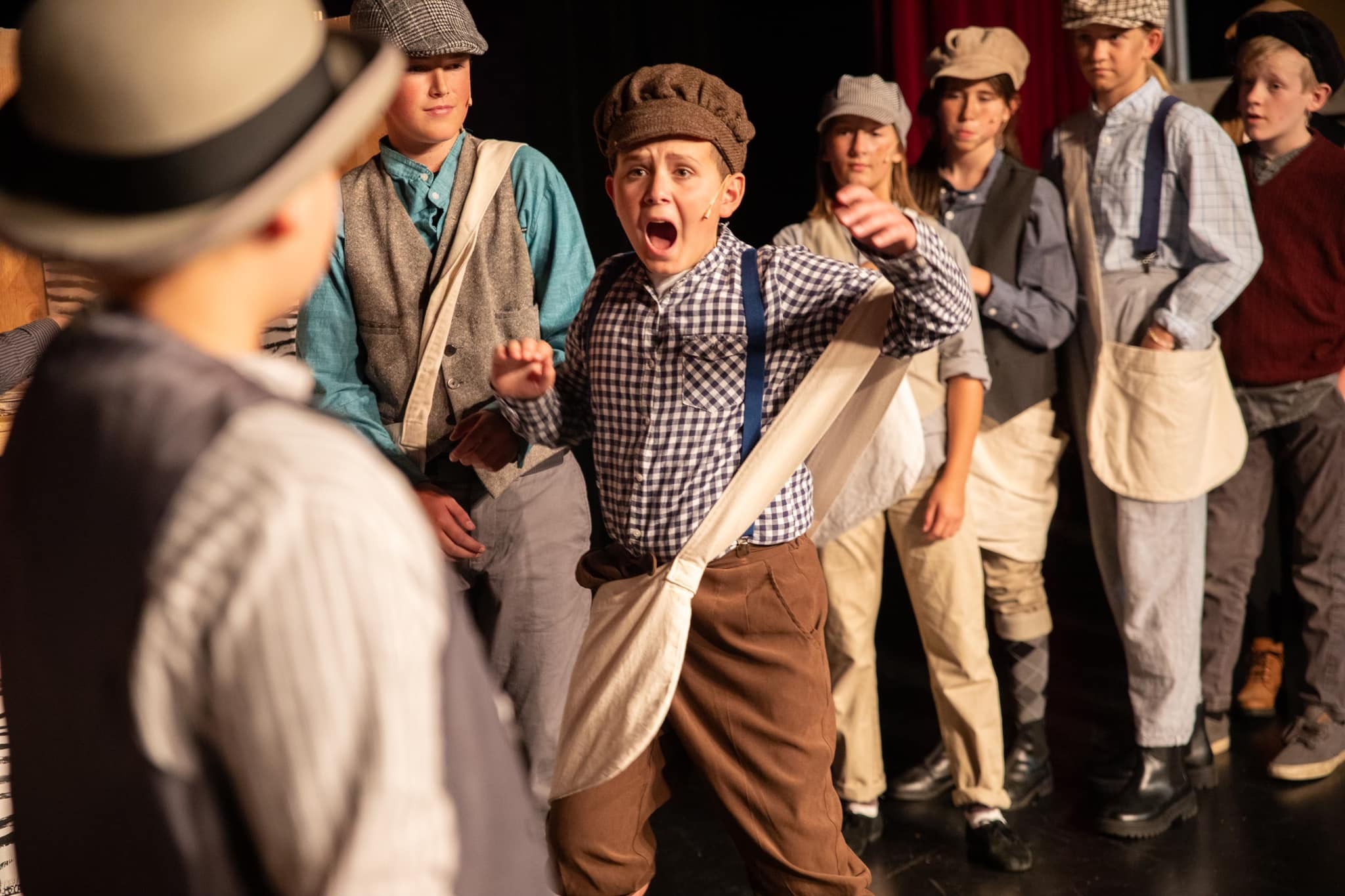 photo from Newsies of boy  in early 1900's dress with mouth open and newspaper bag around should with other kids behind him in similar fashion