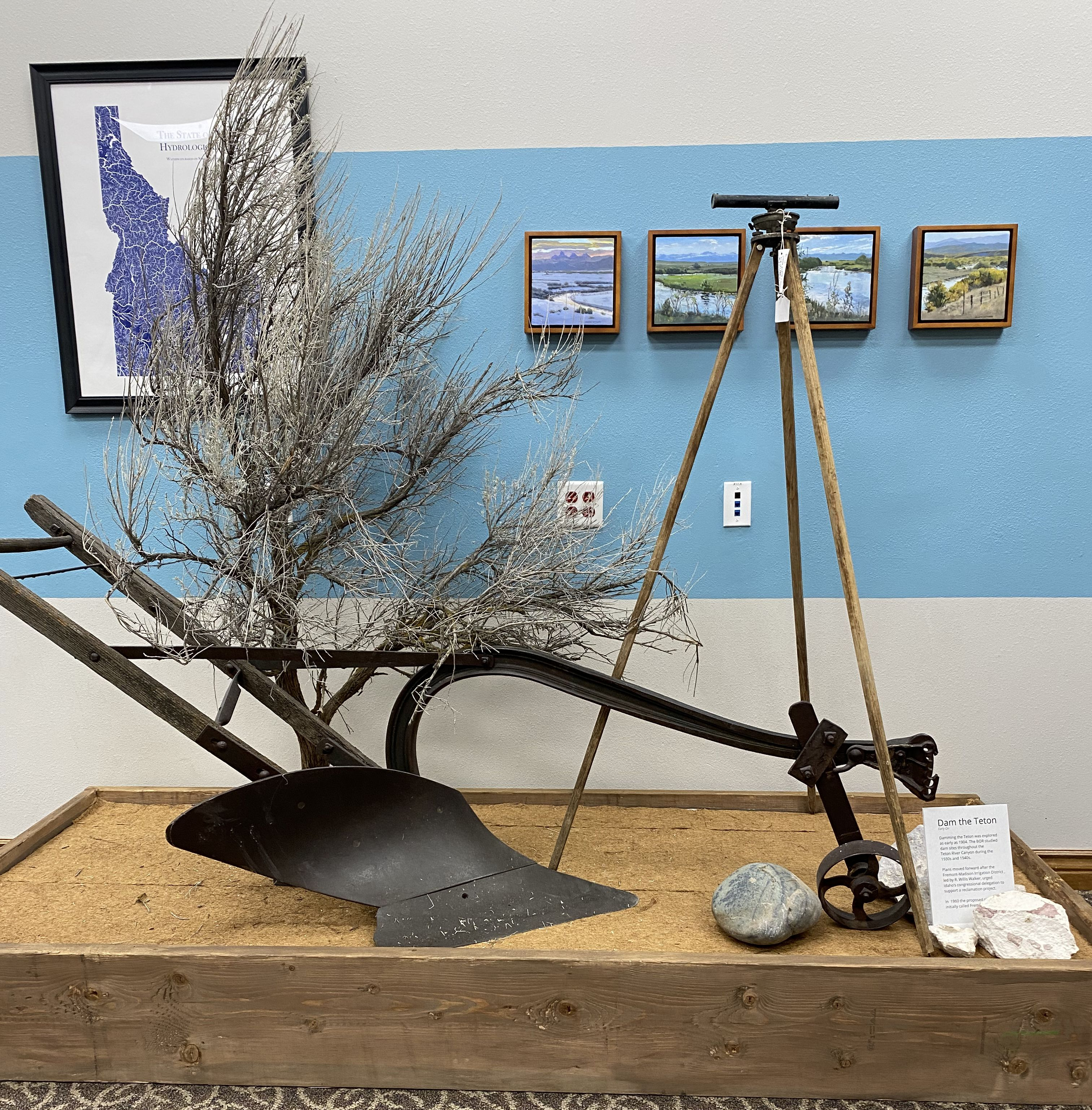 Display of old farm tools on wooden platform against blue wall with framed landscape pictures.