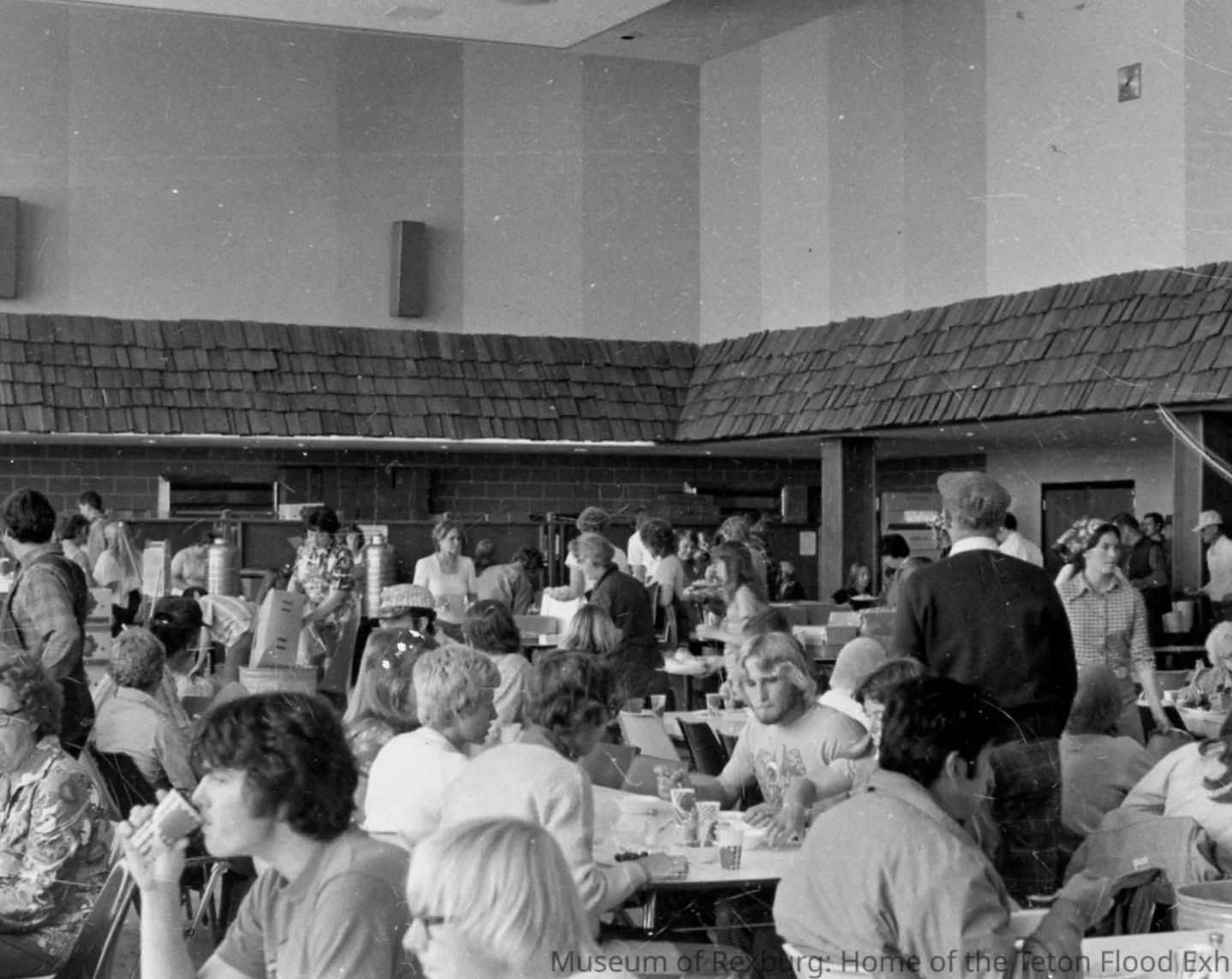 Black-and-white photo of a crowded cafeteria with tables and chairs, people seated and standing, and an open ceiling.