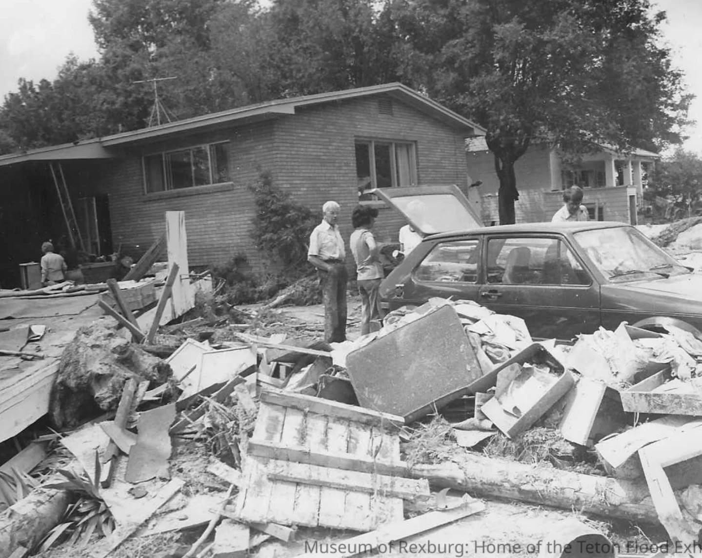 Black-and-white photo of a house in ruins with two people standing in front of a wrecked car.