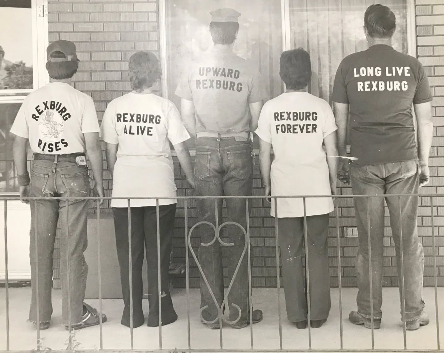 A black-and-white photo of five individuals standing by a railing in front of a brick building.