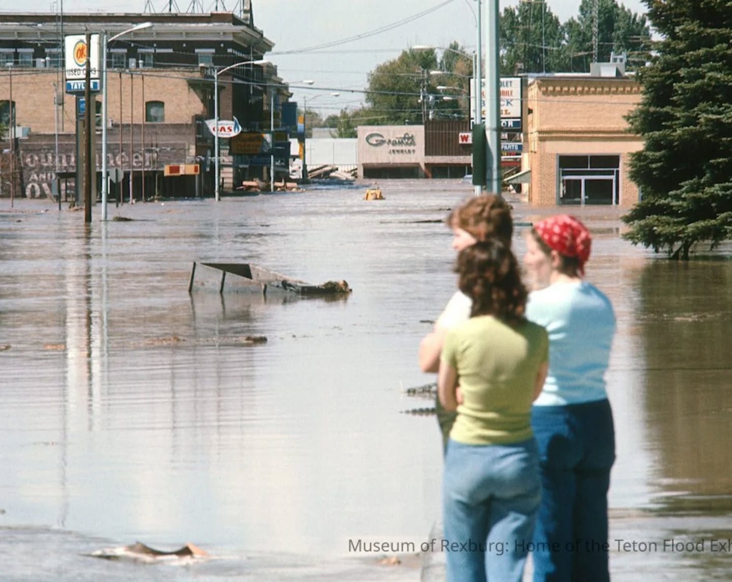 Three people in a flooded street with partially submerged buildings and trees in the background.