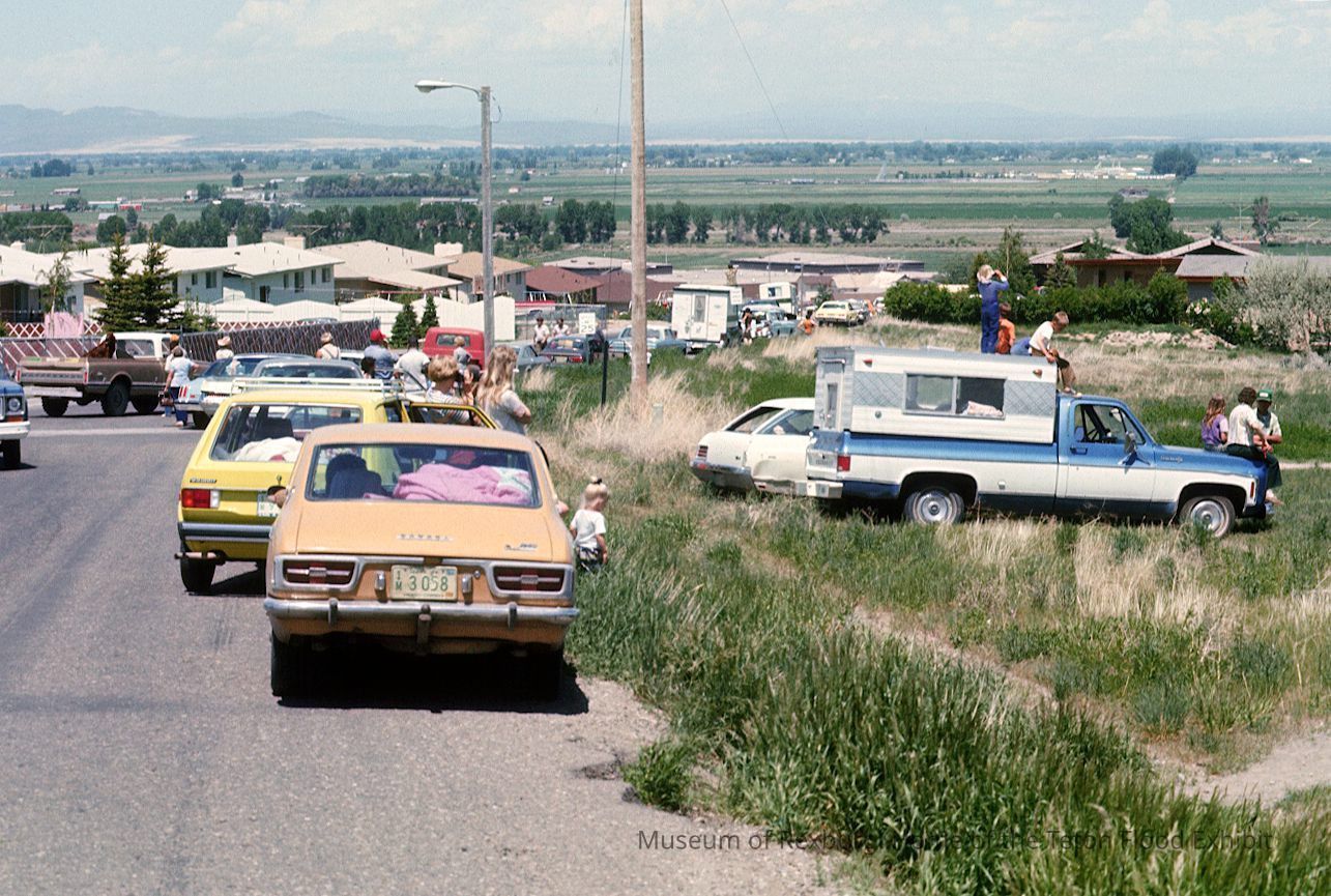 Vehicles parked and driving on a road. People and structures in the distance. Flat landscape.