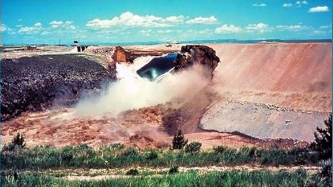 Dirt and debris in an open pit, with a piece of machinery dumping waste. The area is surrounded by grass and has a blue sky.