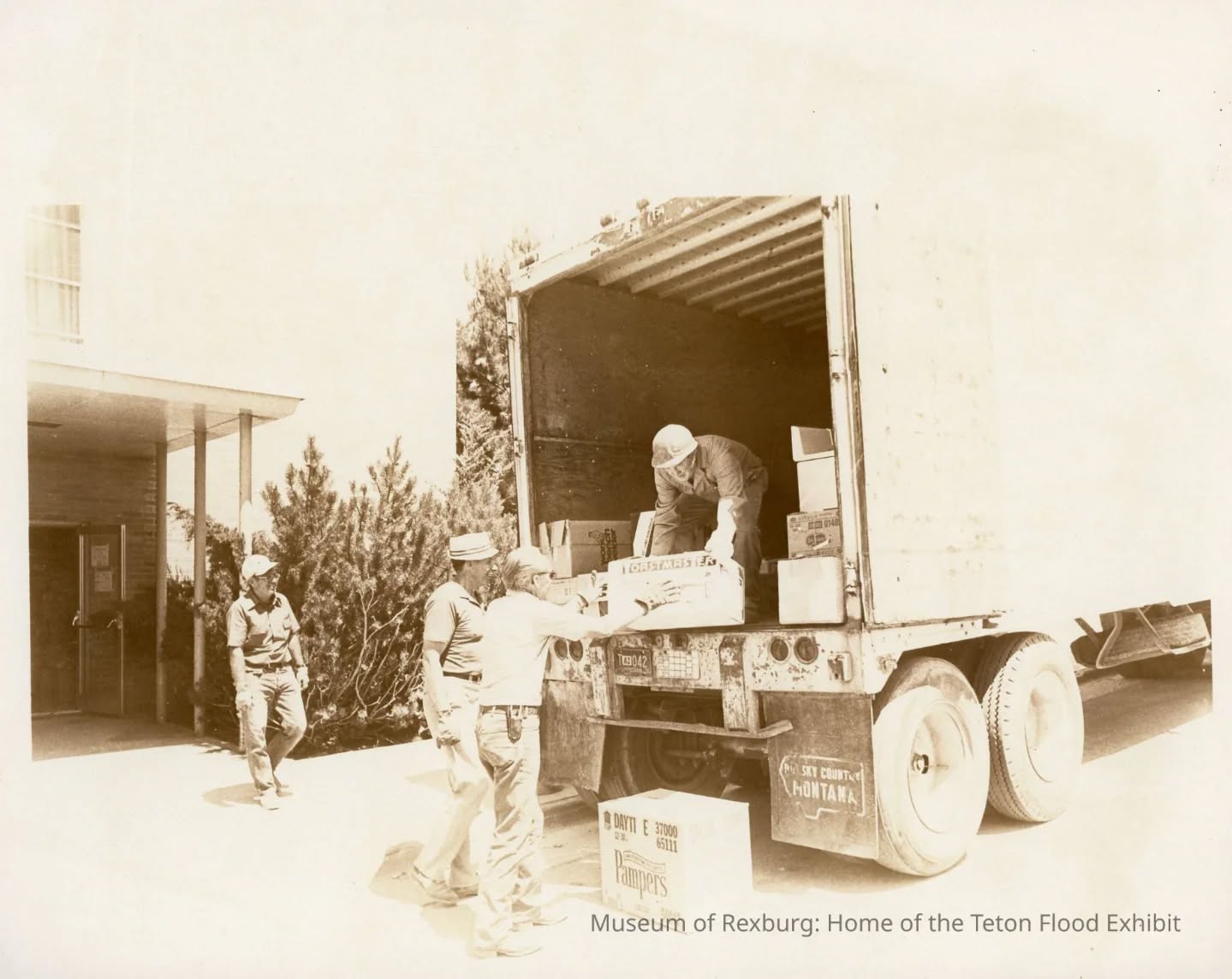 Black-and-white photo of a man unloading boxes from a truck, with two others assisting. Building in background.