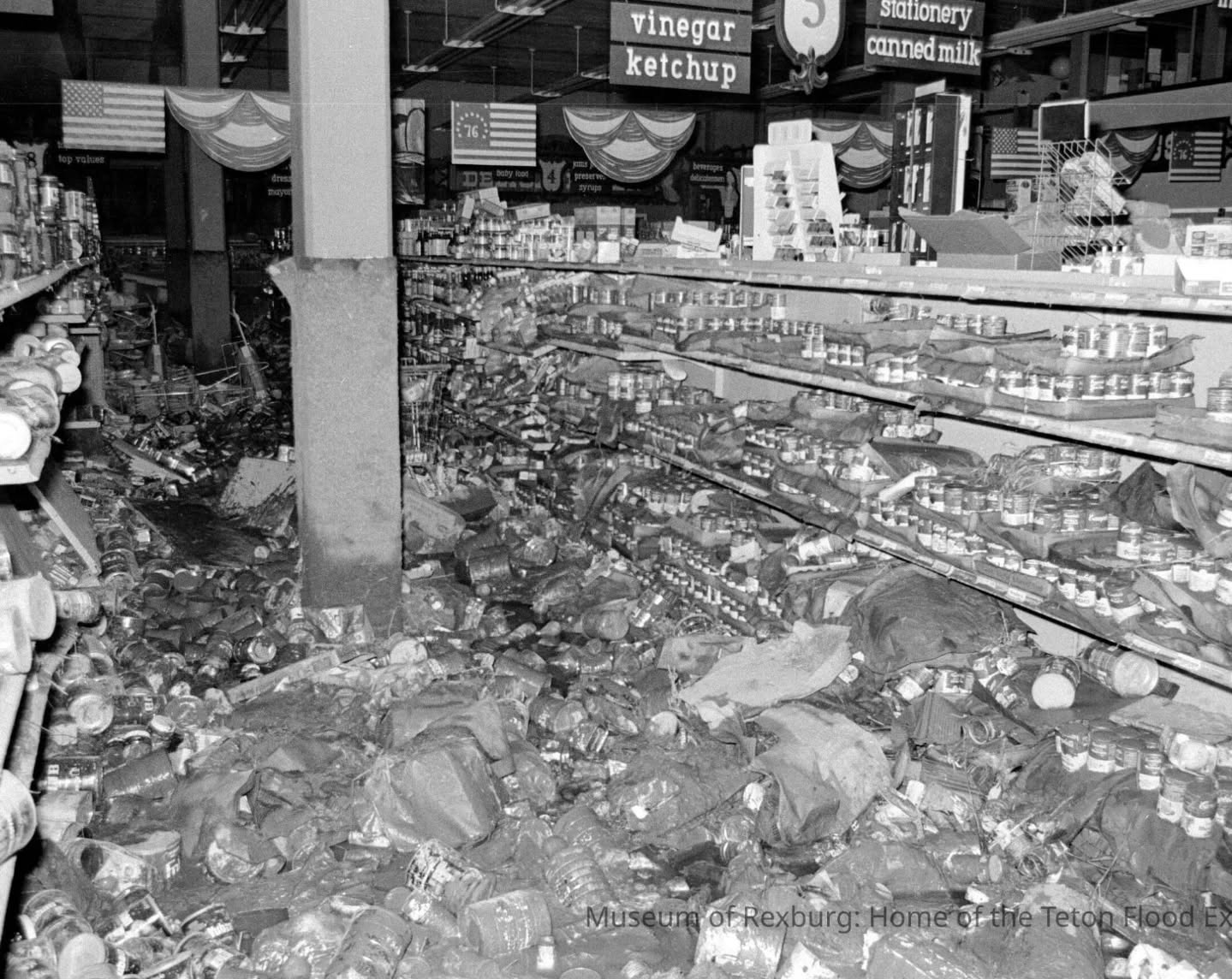 Black-and-white photograph of a store interior. Shelves are collapsed, with products strewn on the floor.