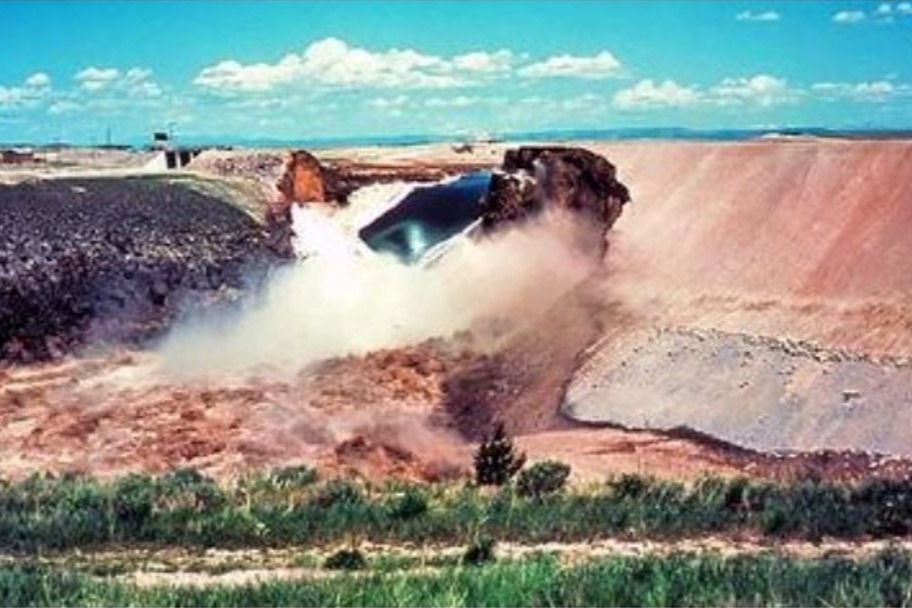 Excavation site with a large machine digging into dirt. Dirt and water spraying. Open area with grassy ground and a blue sky.
