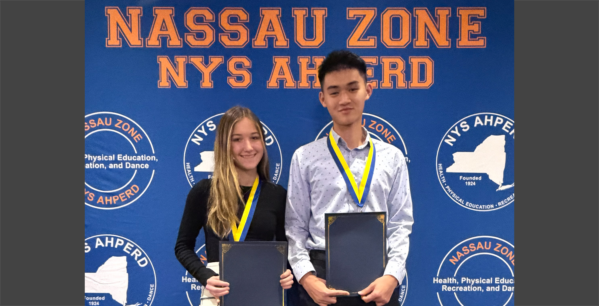Two young individuals, wearing medals and holding certificates, stand smiling in front of a Nassau Zone NYS AHPERD banner at an awards event.