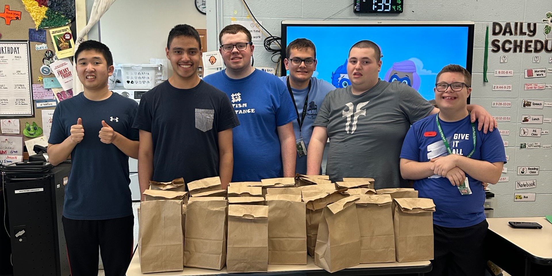 Six smiling young men stand behind a table filled with paper bags. They are in a classroom setting with educational posters and a daily schedule on the wall.