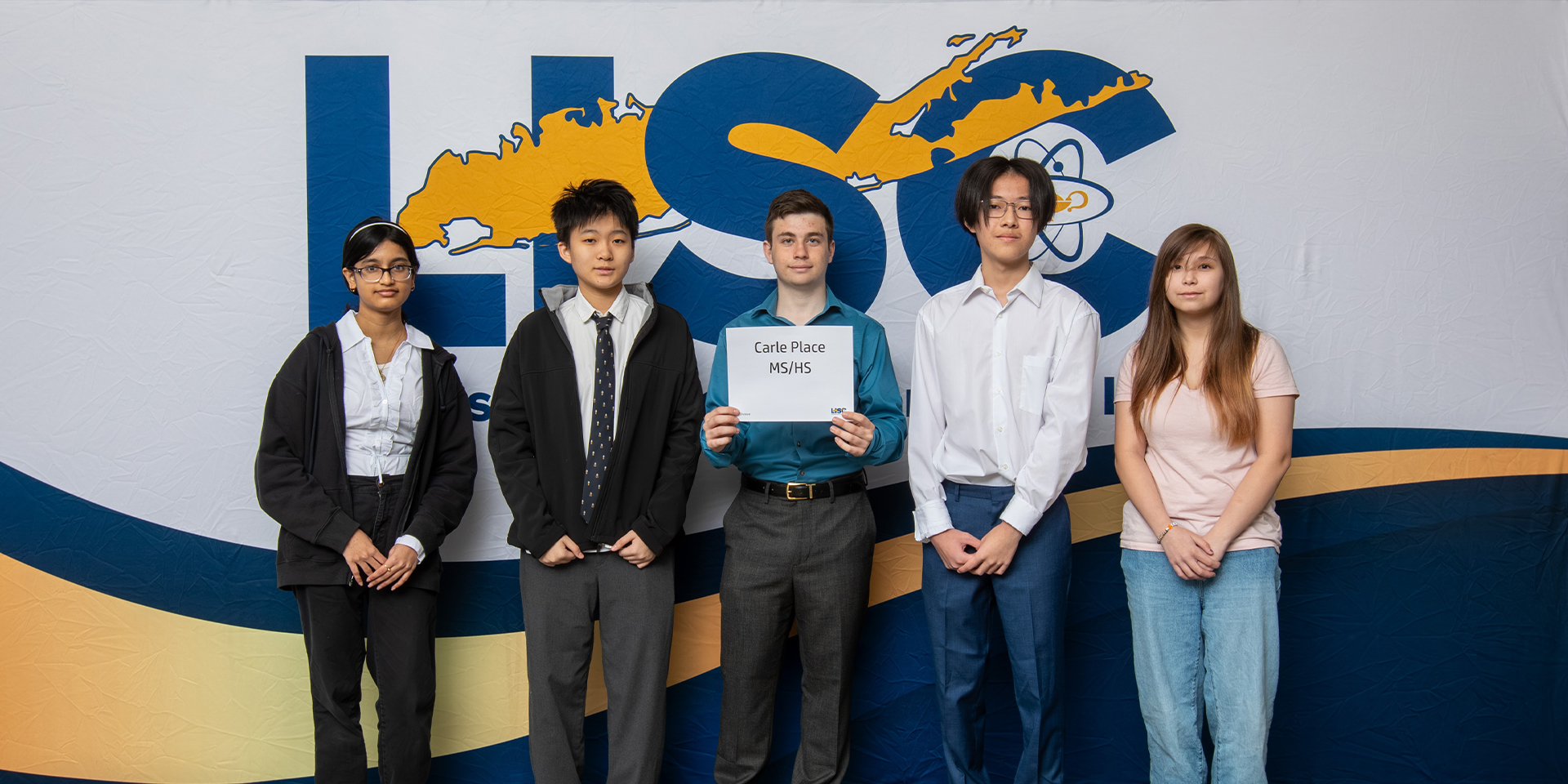 A group of five young people poses in front of an "LISC" banner. The person in the center holds a sign reading "Carle Place MS/HS." They appear focused and proud.