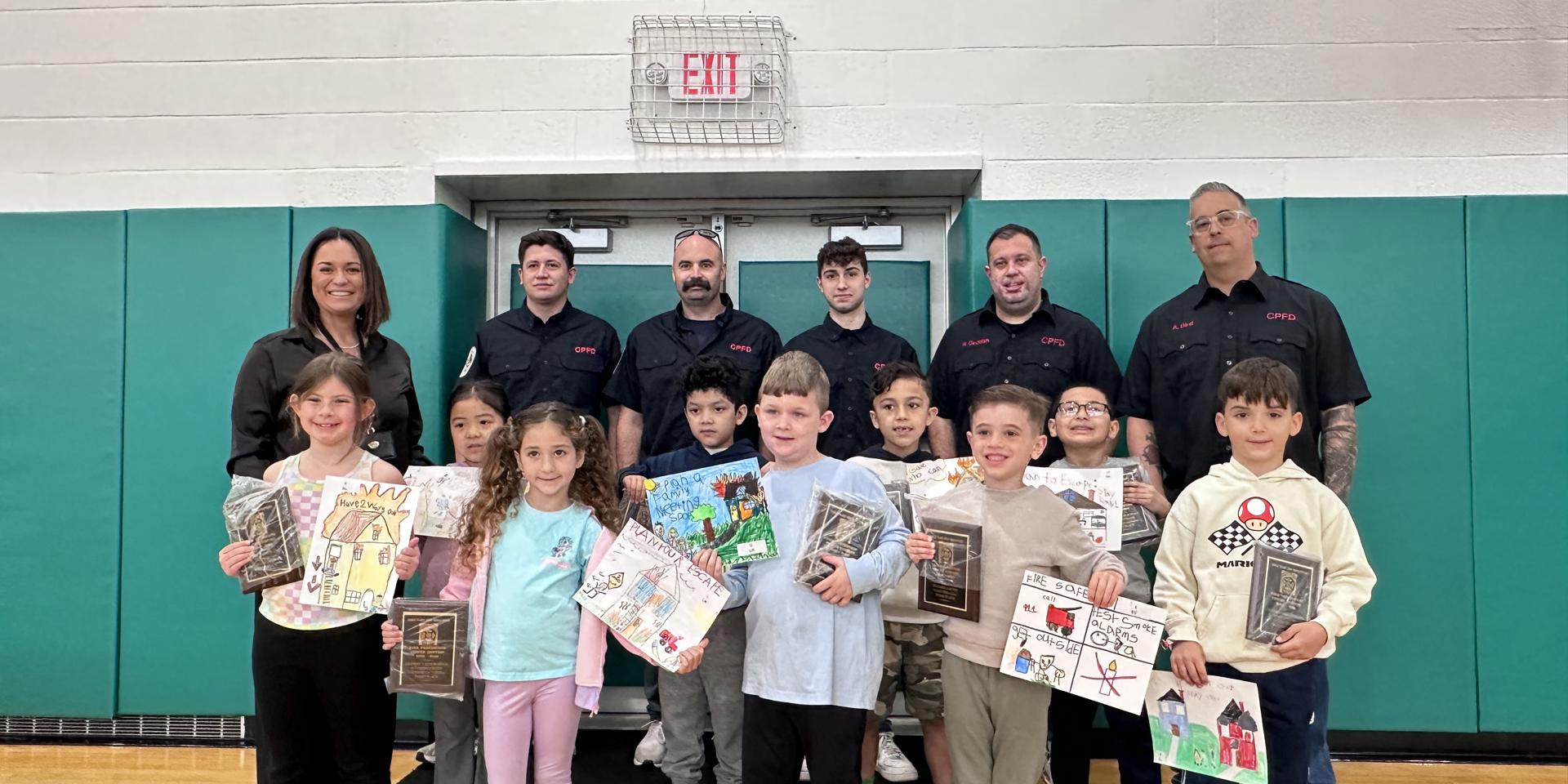Group photo of kids and mentors in a gym, showcasing children's artwork and awards, promoting community engagement and education.