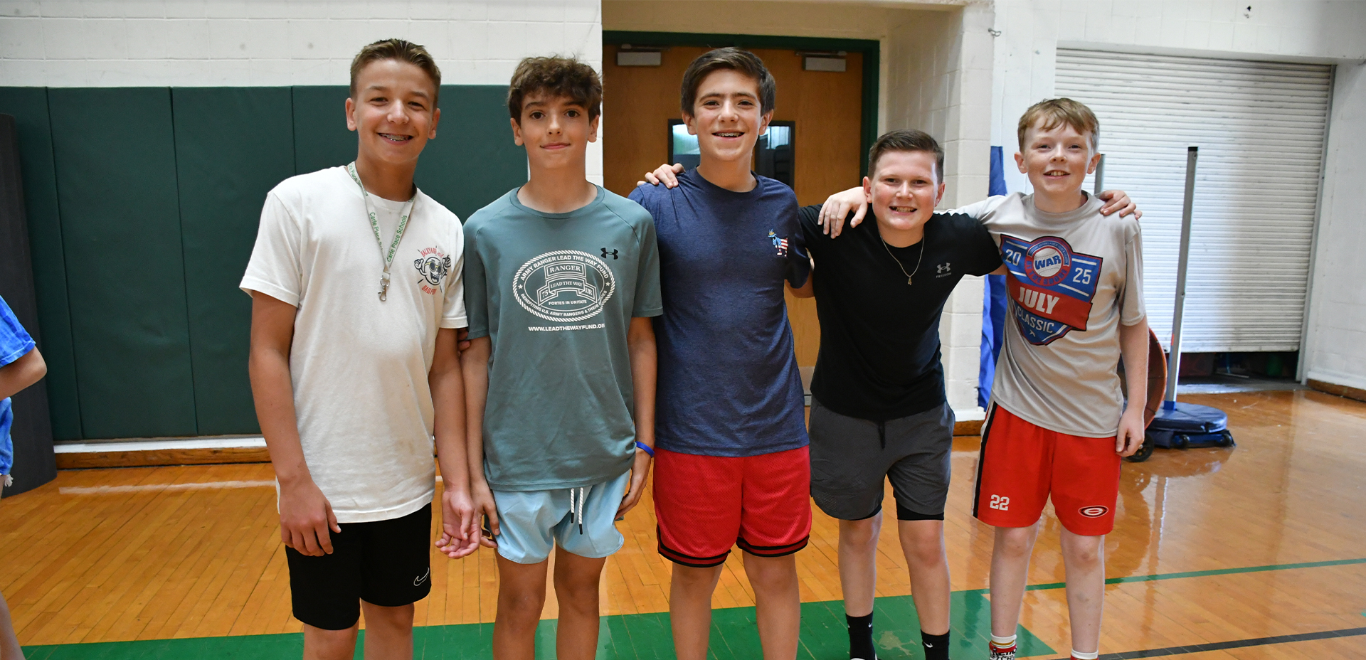 Group of five boys smiling together in a gymnasium, dressed in casual sportswear, showcasing friendship and active lifestyle.