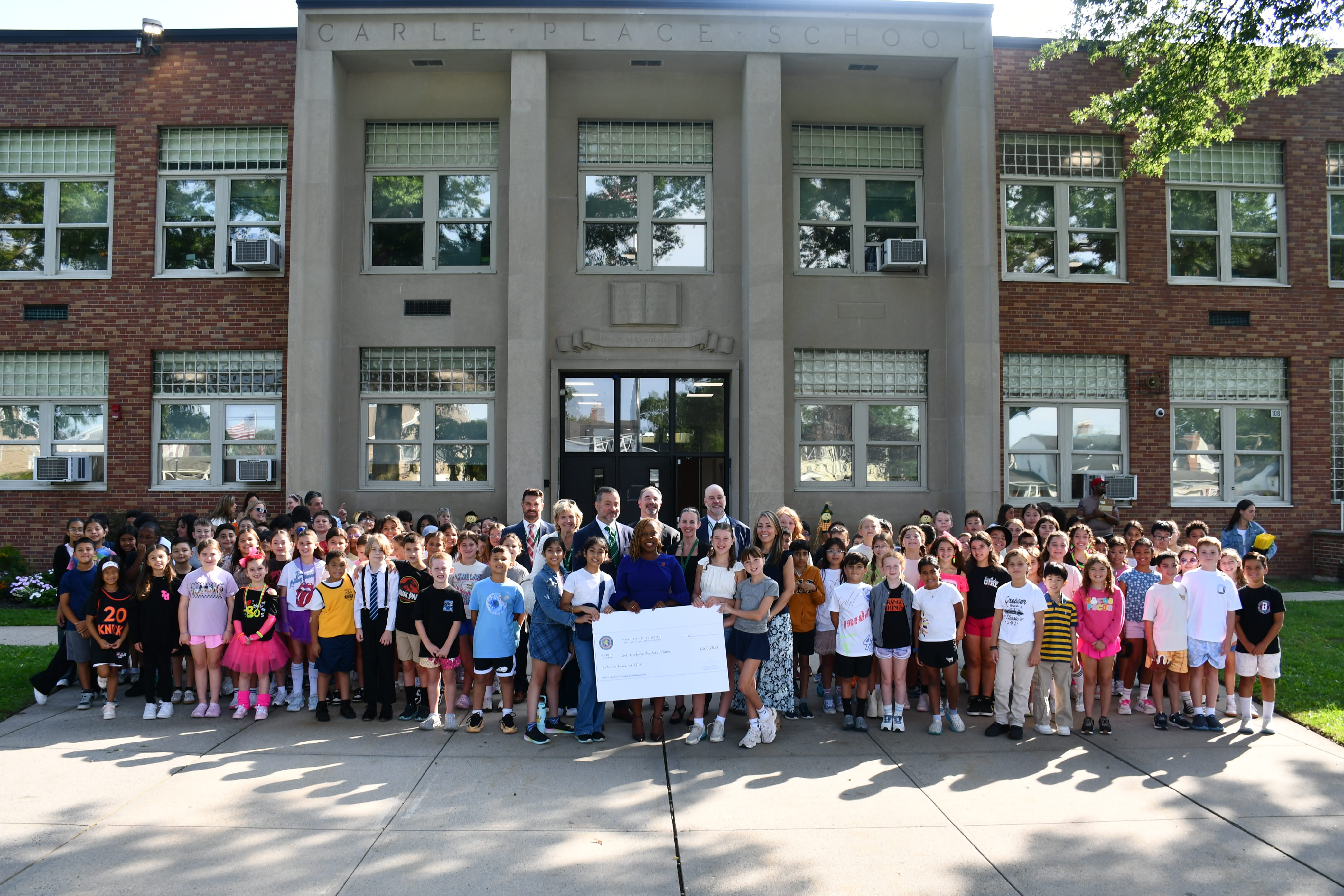 A group of people stands in front of Carle Place School, holding a large check.