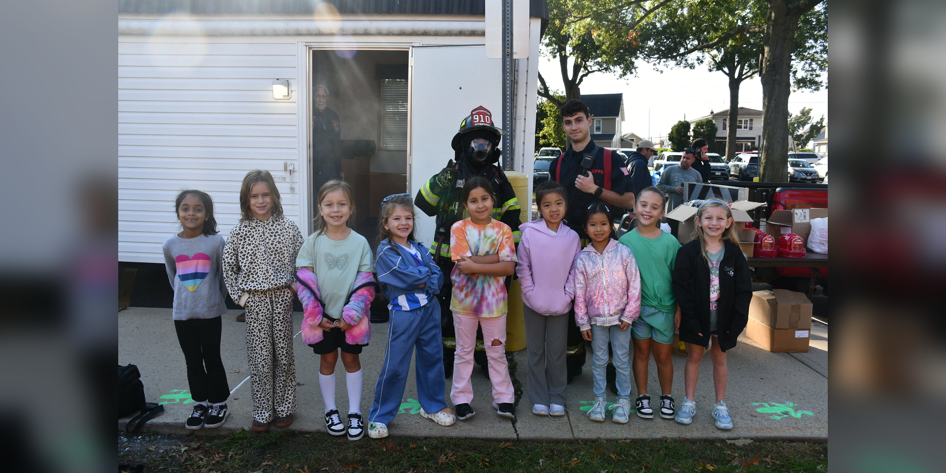 A group of children poses with a firefighter outside a trailer and parked vehicles.