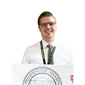 Andy Boone Standing with a yard sign for Presidential Award in front of the Gretna Public Schools logo on the wall