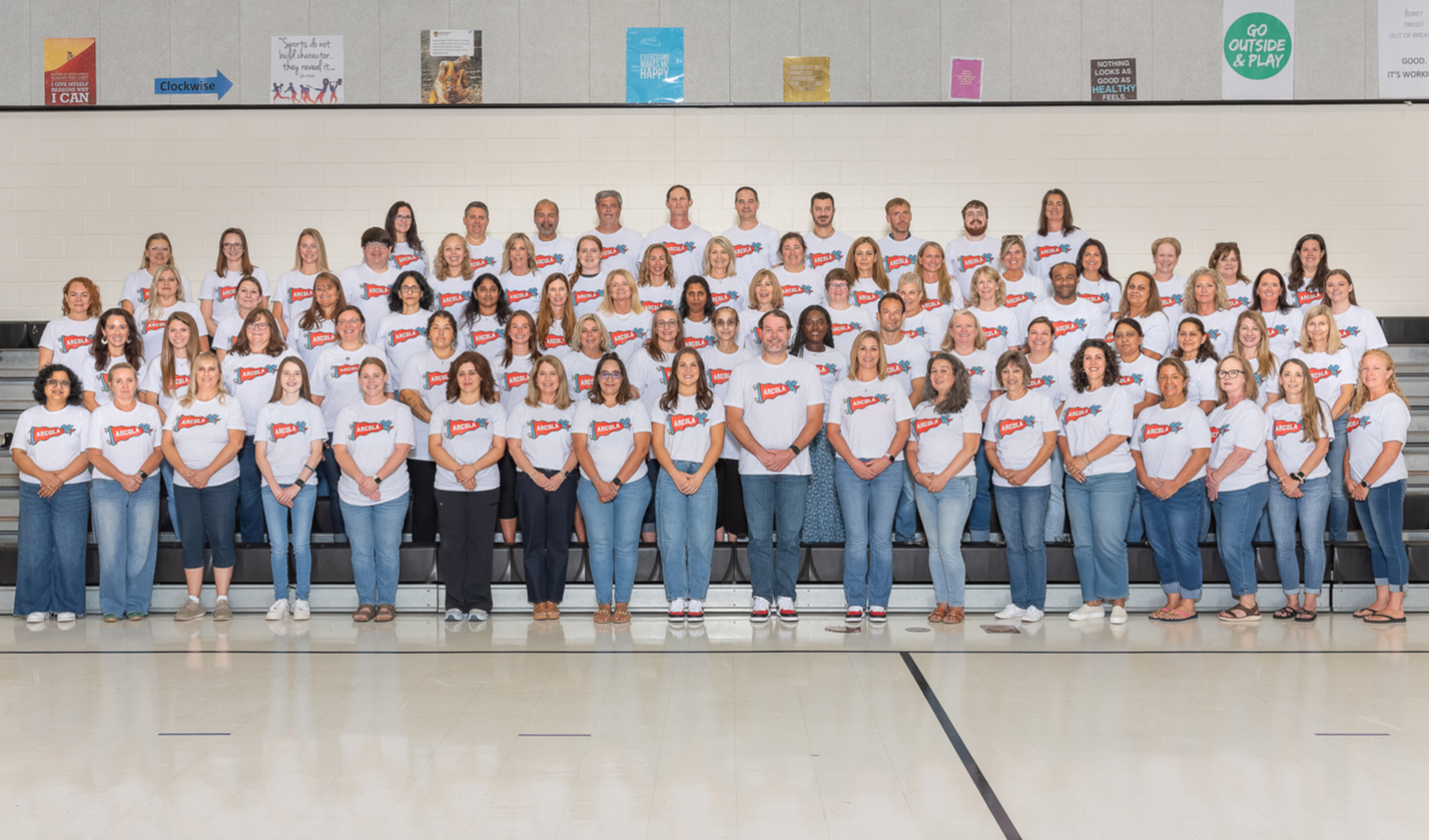 Group of adults and students in white shirts and blue jeans pose on a bleacher.