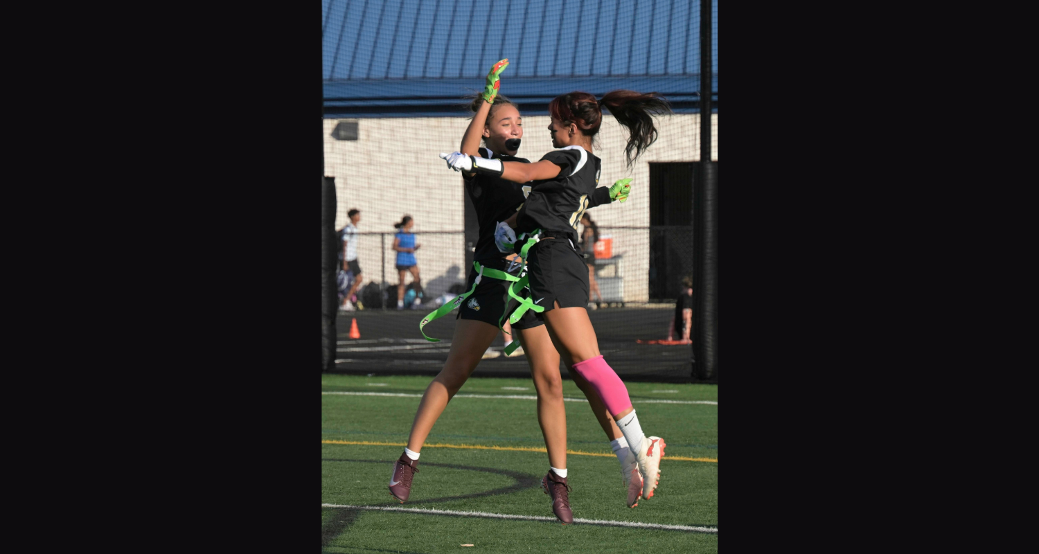 Two women playing a game on a field. One is jumping while holding a ball, and the other supports her.