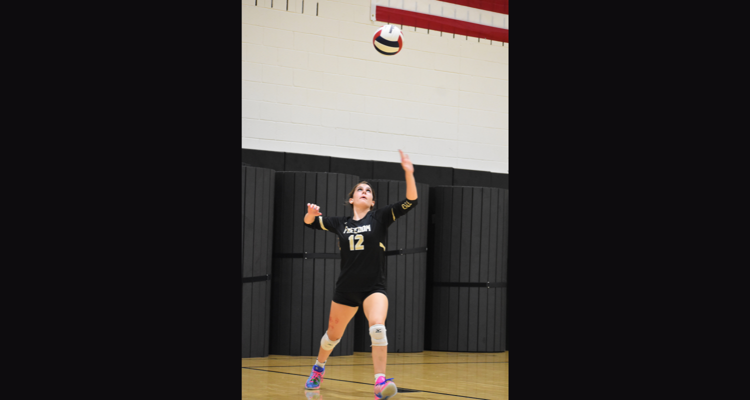 A person in a black jersey with number 12, serving a volleyball in a gymnasium.