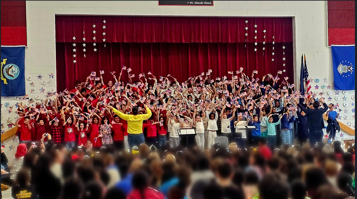 A crowd gathers, many holding flags, in a large room with a stage featuring a red curtain.