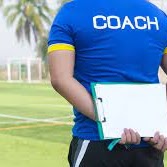 Coach with blue tshirt on standing back facing with clipboard behind his back