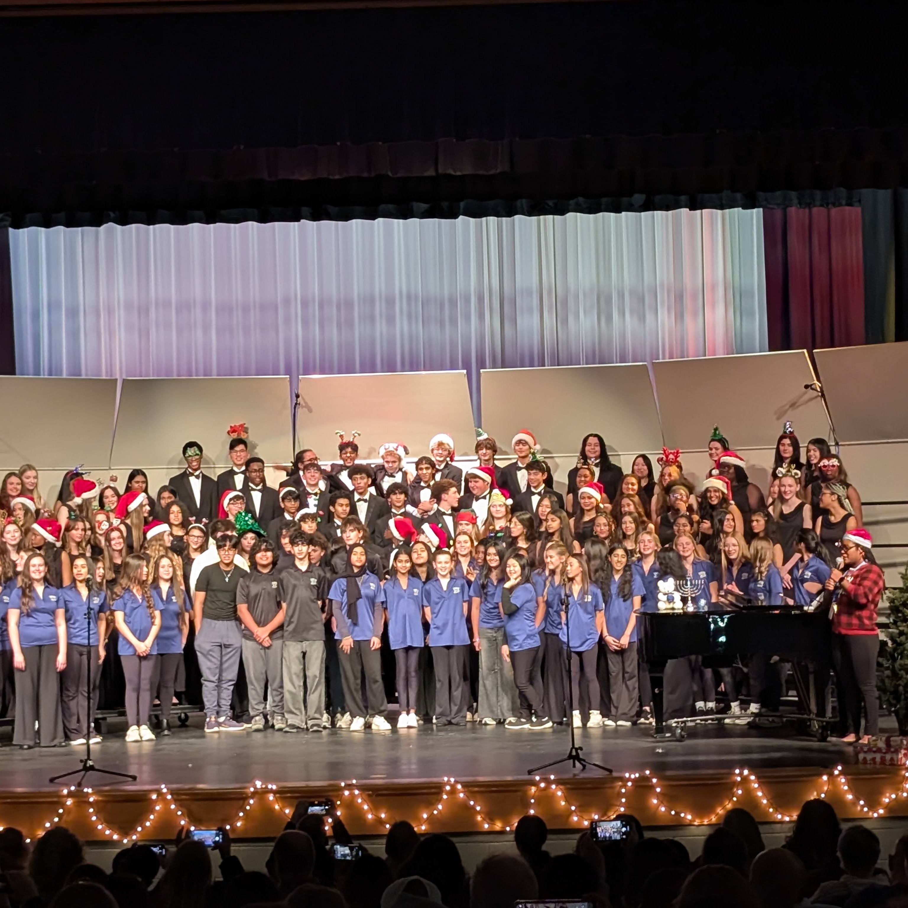 A large student choir stands on an auditorium stage during a holiday concert, with many members wearing Santa hats.