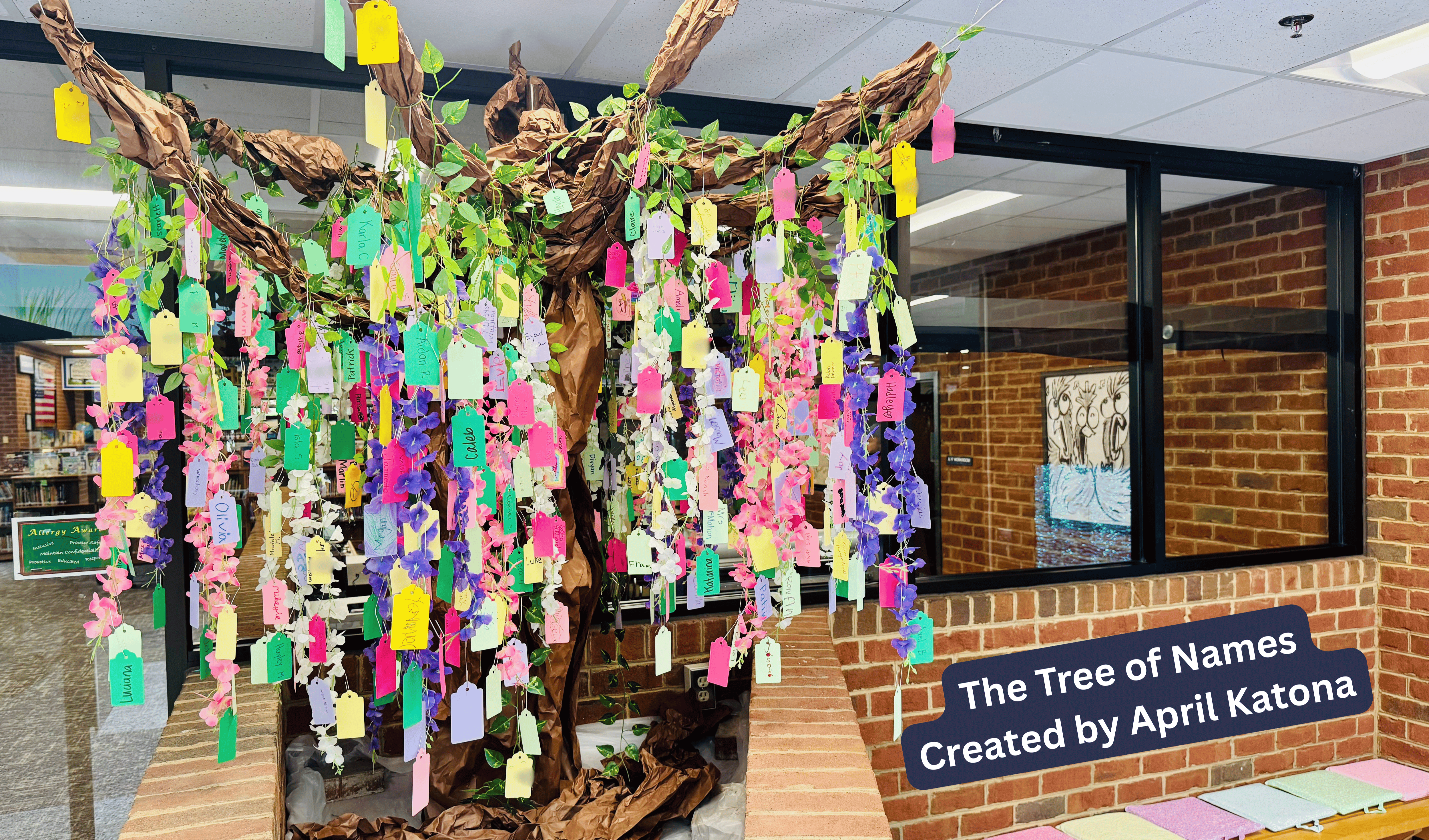 Colorful tags hang from a tree trunk in a room with brick walls. A sign reads, "The Tree of Names Created by April Katona."