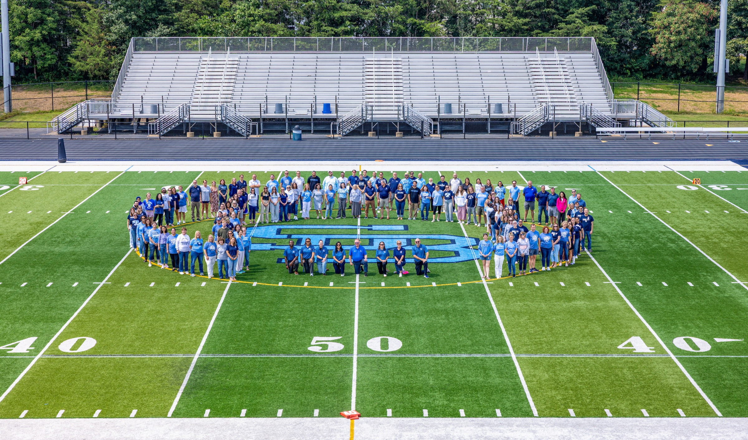 A large group of faculty and staff members wearing blue and white clothing assemble on an athletic turf field, arranged in a wide formation surrounding a large blue "SB" logo painted at center field.