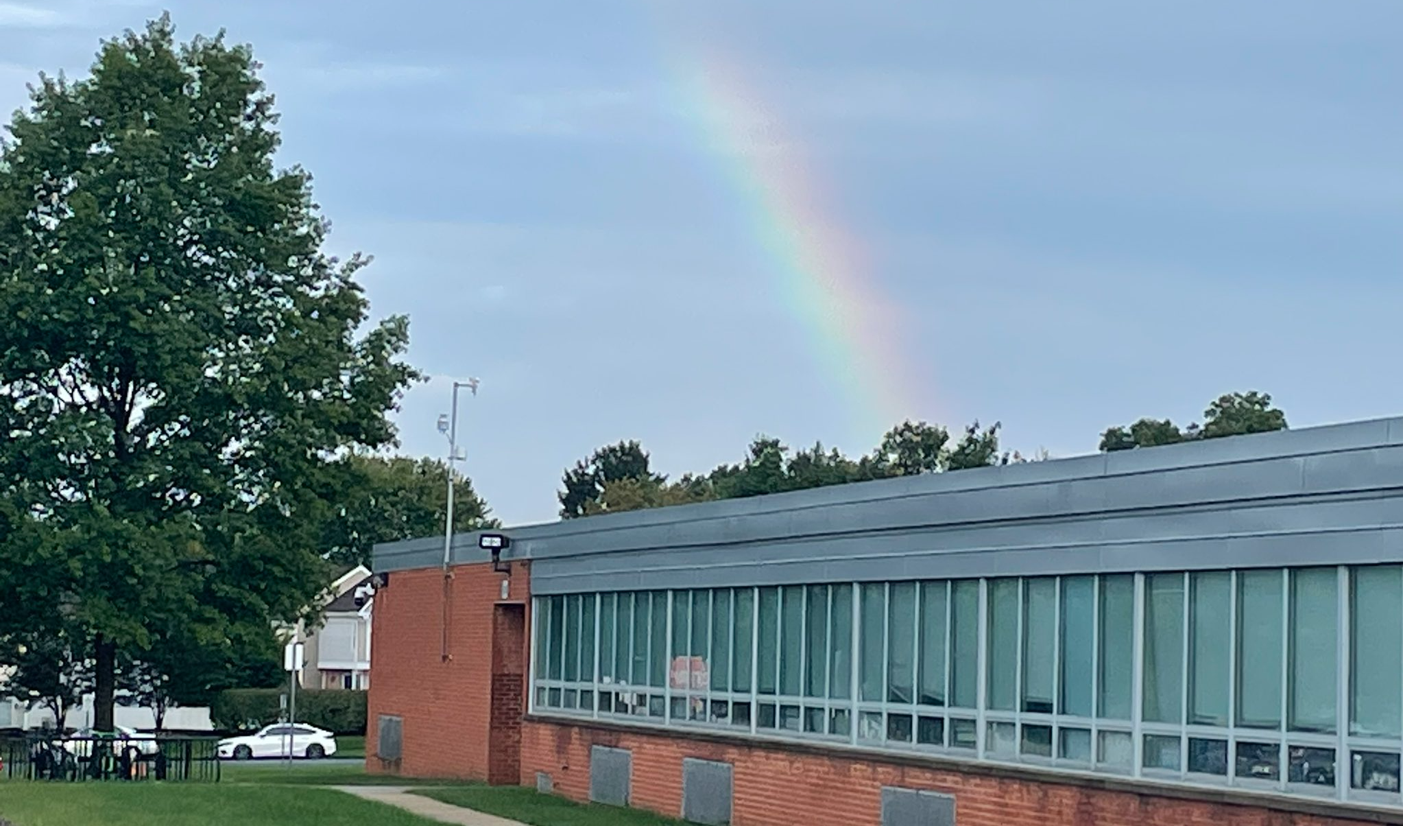 A photograph of a partial **rainbow** stretching across a soft blue and grey sky. The rainbow appears over the flat roof of a single-story brick school building with a long row of large windows. To the left of the building, there is a green lawn with a large, leafy green tree and a white car parked in the background.