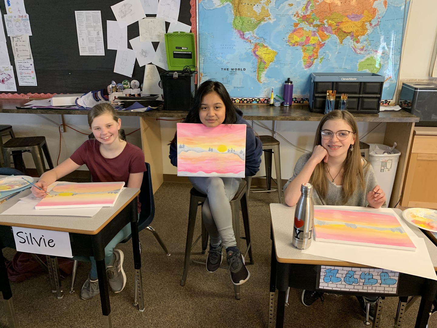 three students inside a classroom each holding their art activities