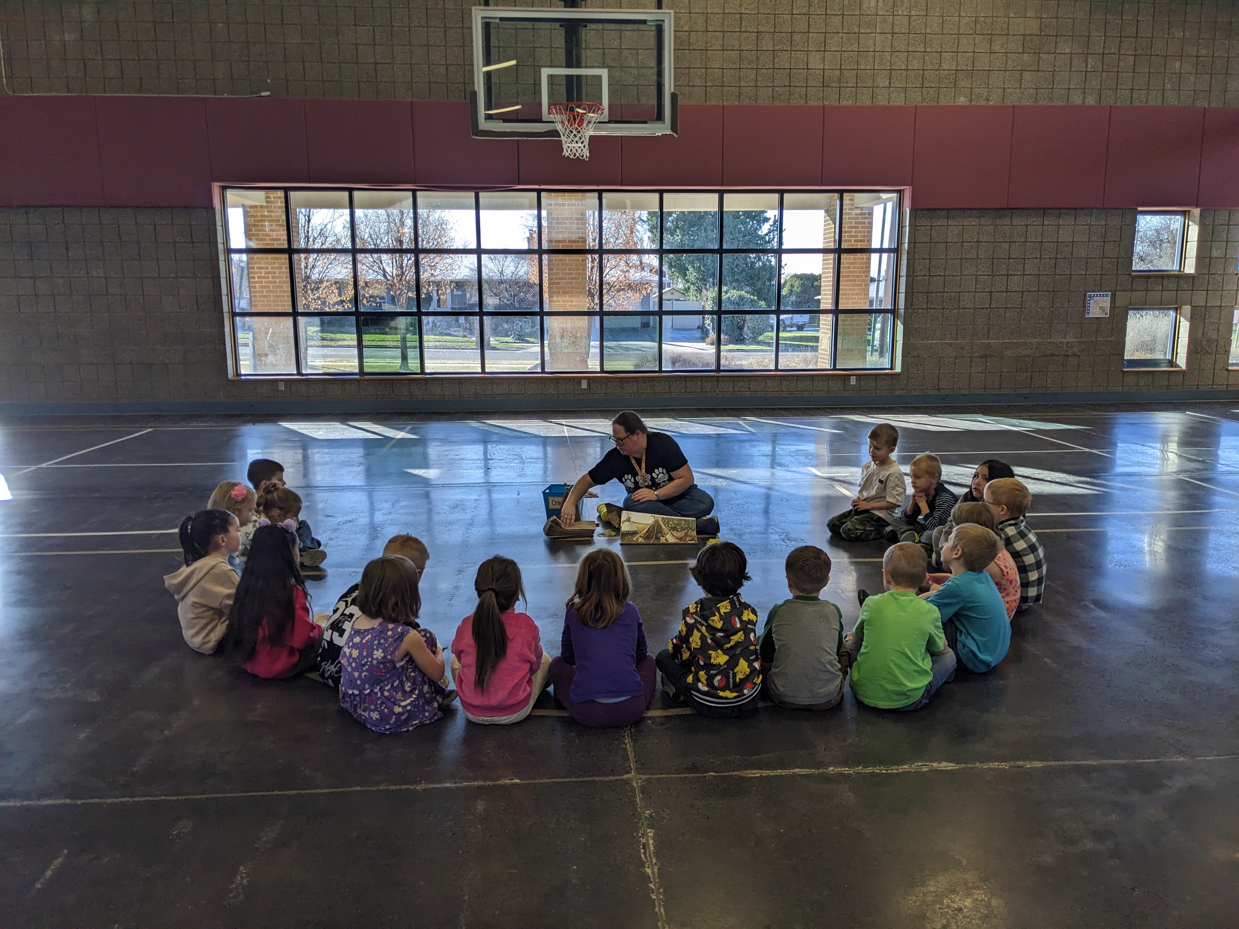 A group of kindergarteners sitting on the gym floor, surrounding a teacher.