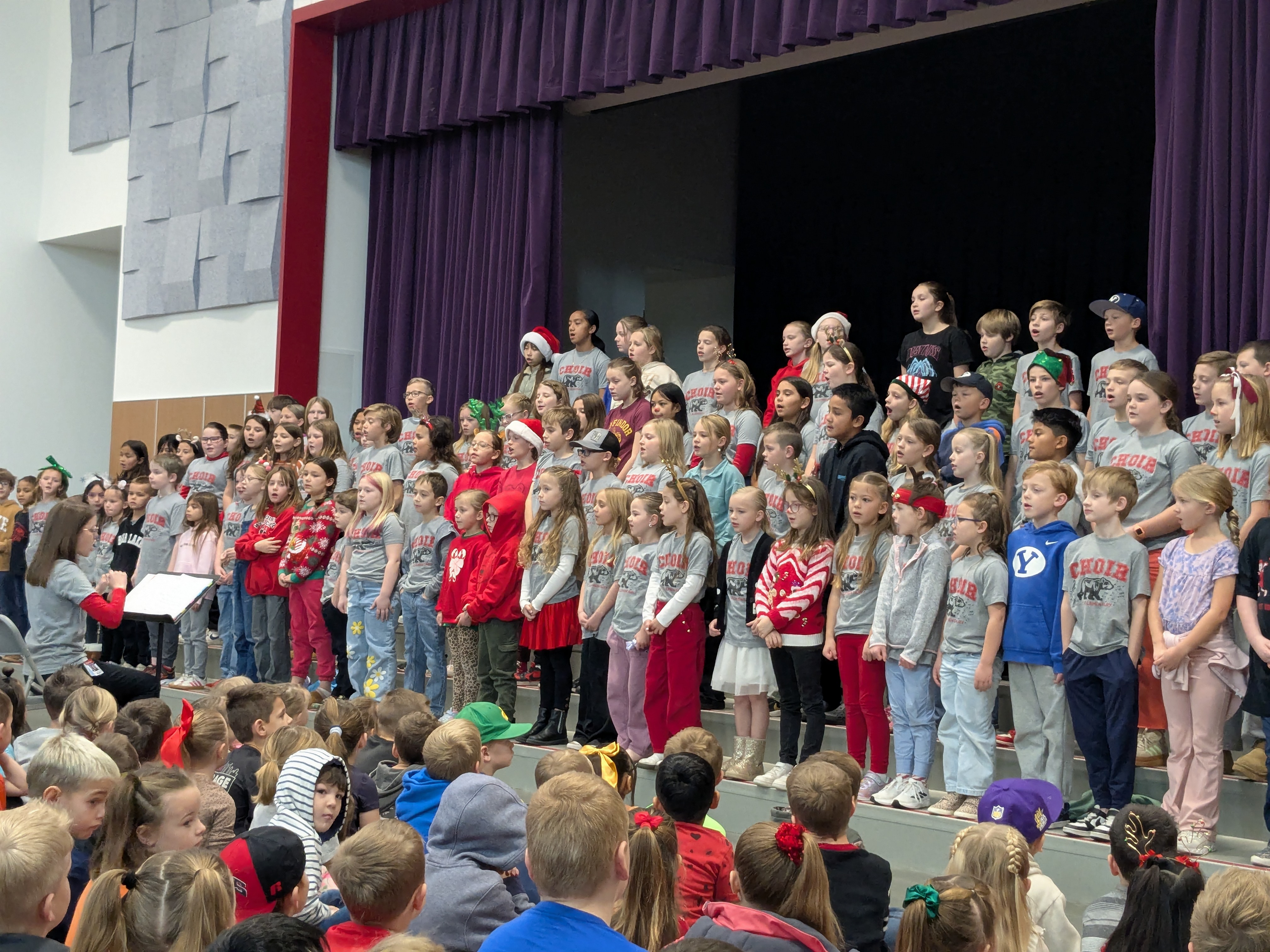 West Bountiful Elementary Choir performing at a school assembly. Students are standing on the stage with their conductor leading them.