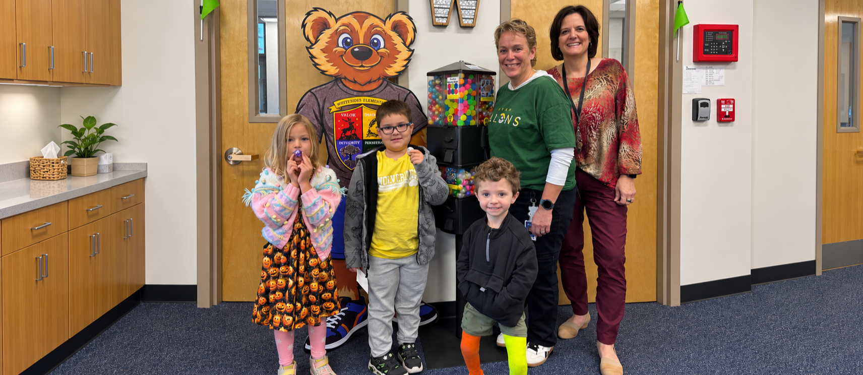 Three students and two school principals pose in front of Winston the wolverine, the school mascot and a candy vending machine.